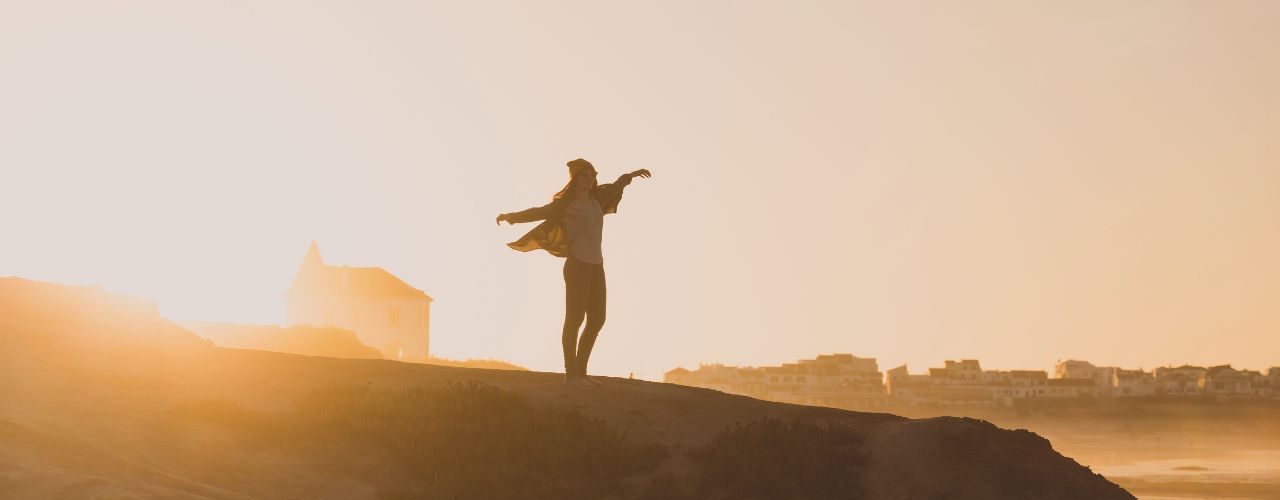 Woman standing on a mountain with arms outstretched, symbolizing blind faith, trust, personal transformation, and embracing uncertainty.